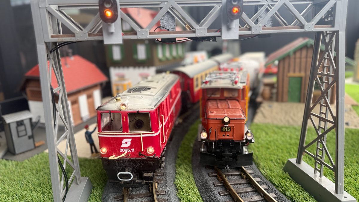 Two G scale locomotives side by side under a signal gantry